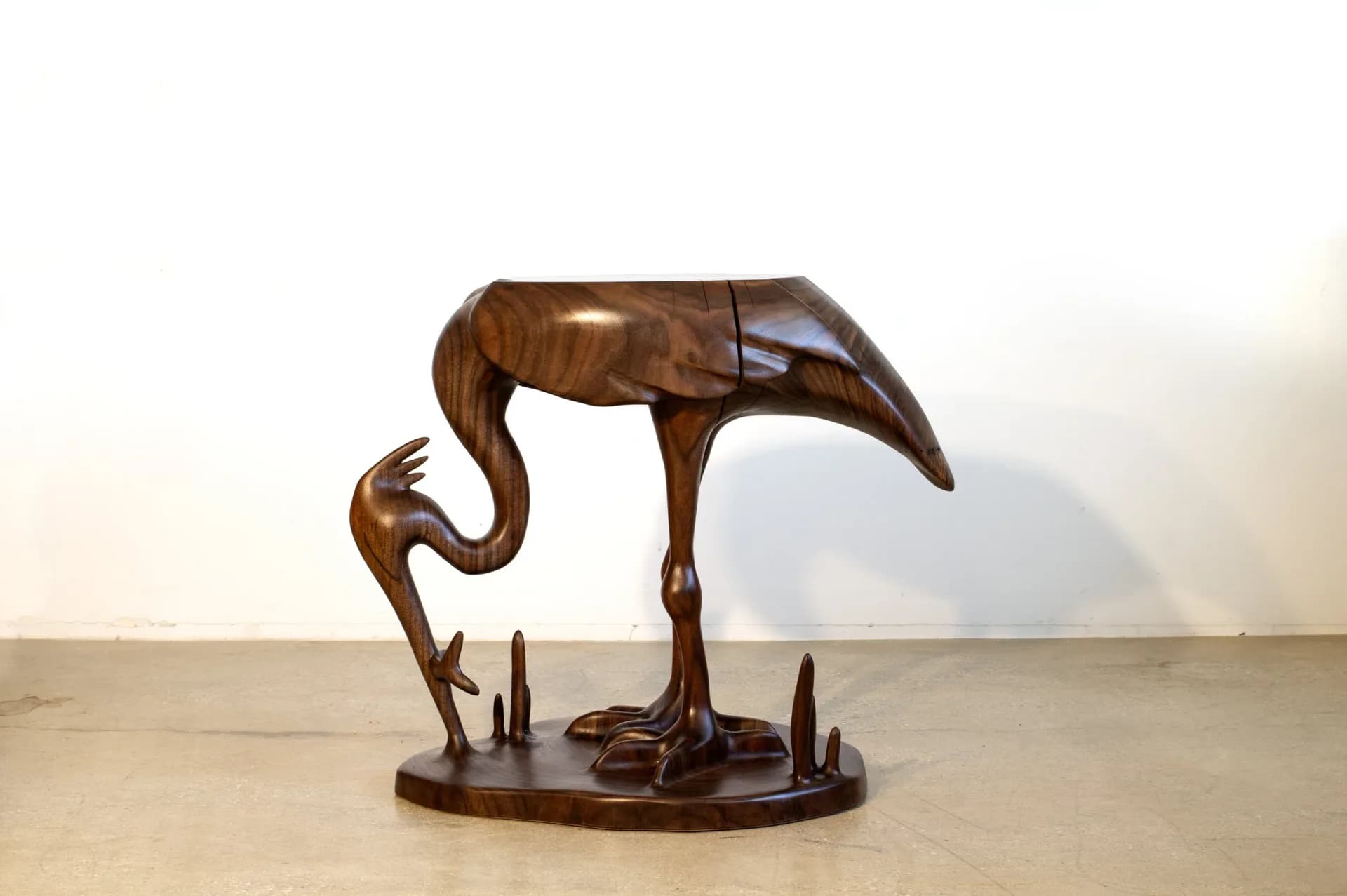 Wooden sculptural bird figure with curved neck and legs on a dark base, displayed in a minimalist gallery space by Aaron Poritz.