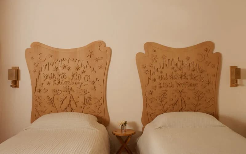 Bedroom with two twin beds featuring carved wooden headboards with decorative relief detailing, white bedding, and a small wooden side table between them by Adam Charlap Hyman.