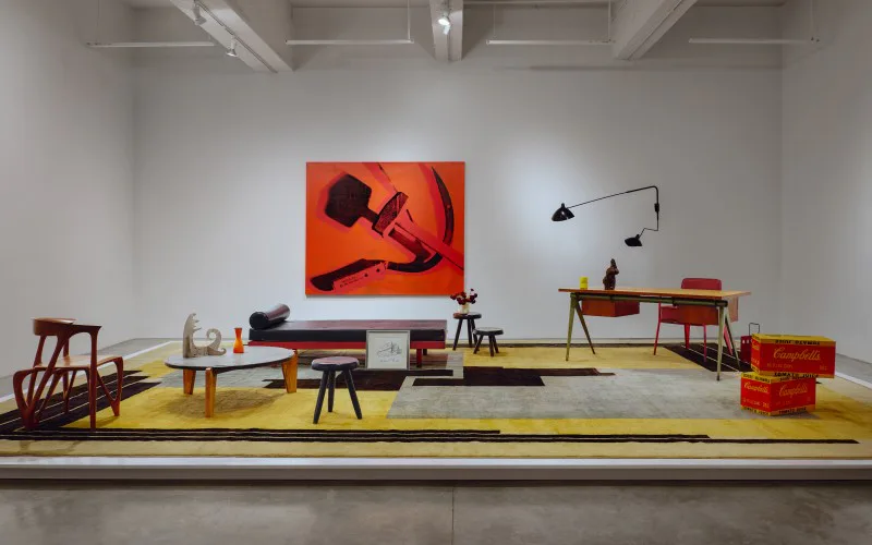 A mid-century modern furniture showroom display with mustard-yellow flooring, wooden chairs, tables, and a wall-mounted sculpture, featuring an orange abstract artwork by Adam Charlap Hyman.