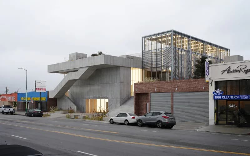 Brutalist concrete structure with angular forms and large windows adjacent to brick industrial building with metal framework, street-level view by Adam Charlap Hyman.