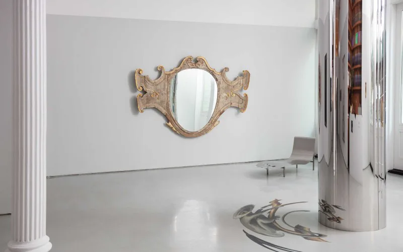 Minimalist gallery space with polished concrete floor, ornate carved wooden mirror on white wall, classical columns, and sculptural elements by Adam Charlap Hyman.