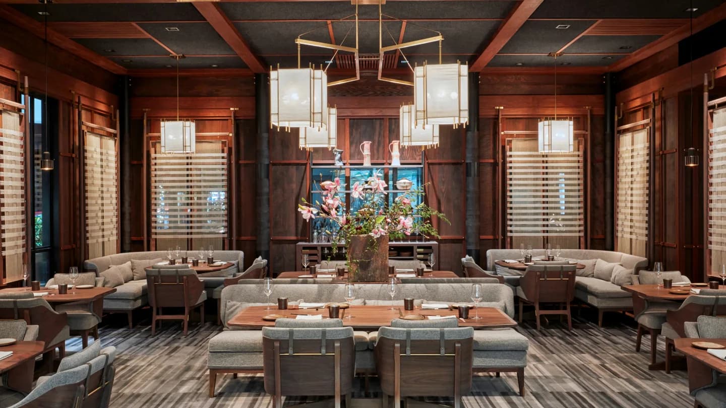 Luxurious restaurant dining room with warm wood tones, symmetrical seating arrangement, modern chandelier, and decorative blue artwork by Adam Farmerie.