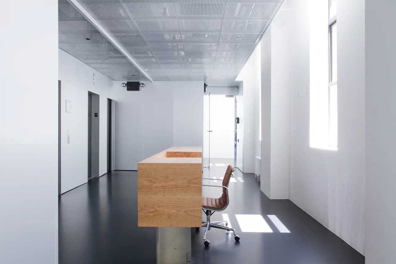 Minimalist office corridor with wooden desk, leather chair, and natural light from clerestory windows, designed by Adam Frampton.