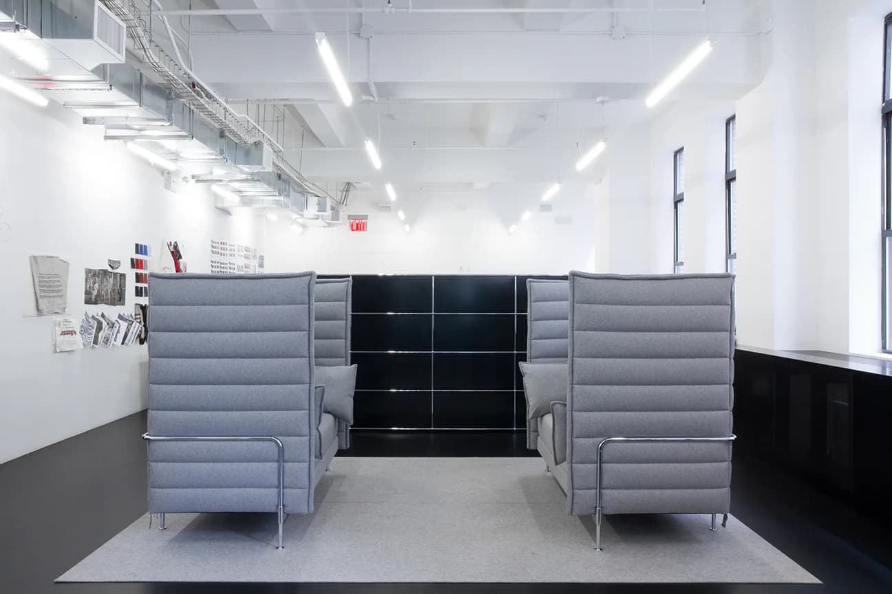 Minimalist interior space with gray upholstered seating elements flanking black shelving units, polished concrete flooring, and bright linear ceiling lights by Adam Frampton.