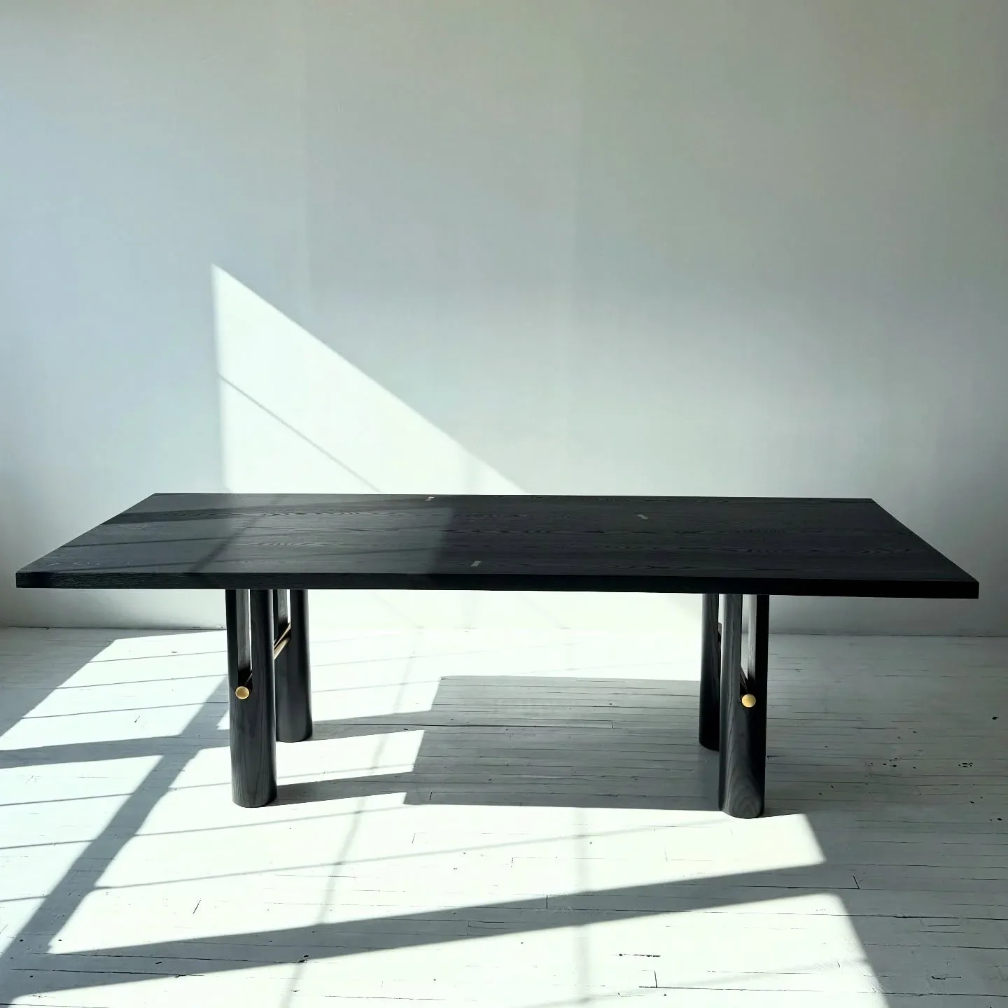 Minimalist black dining table with geometric base in bright white gallery space with dramatic natural light shadows by Adam Johnson.
