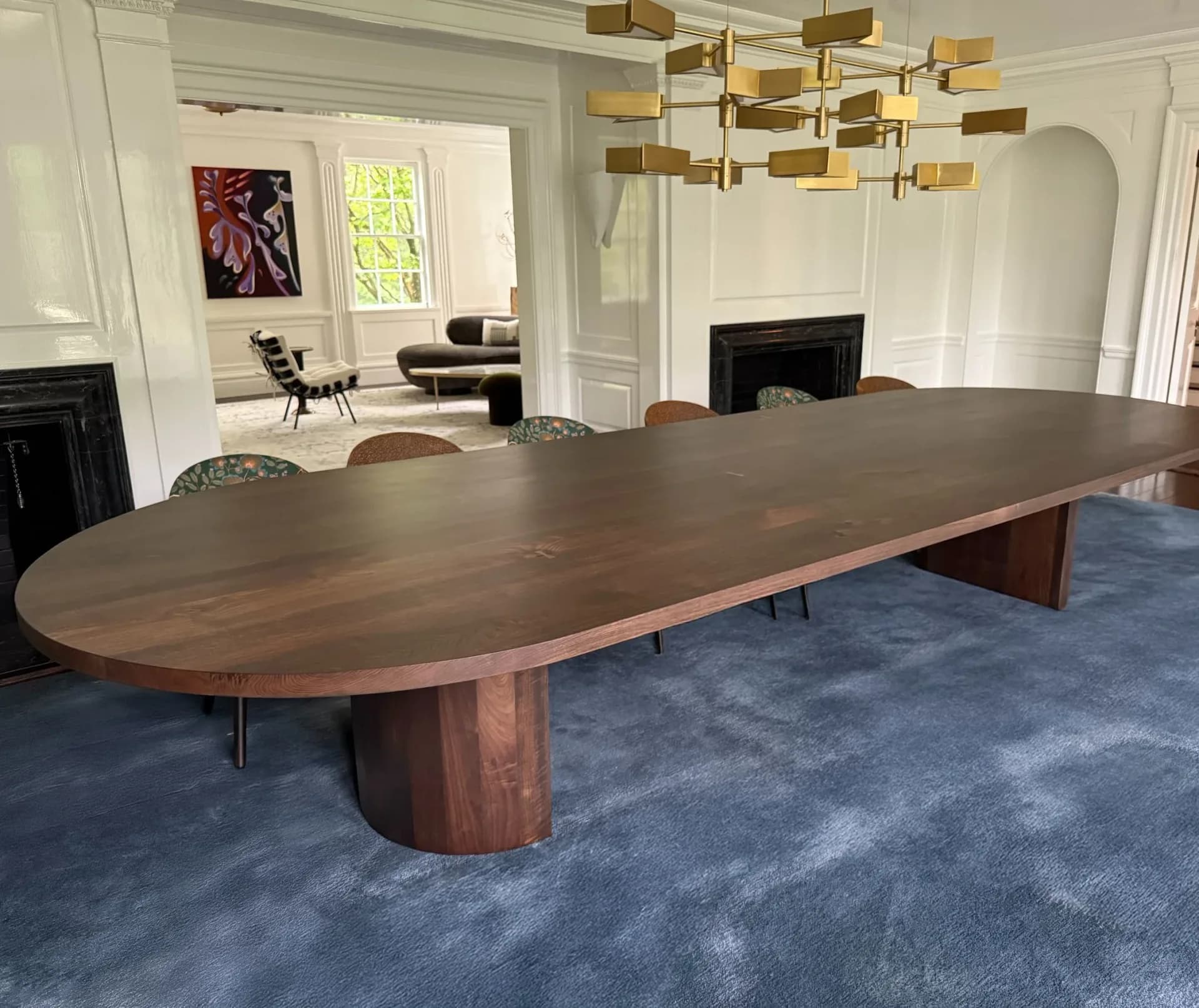 Spacious dining room featuring an elongated walnut table with curved ends and cylindrical base, blue area rug, and modern brass chandelier, by Adam Johnson.