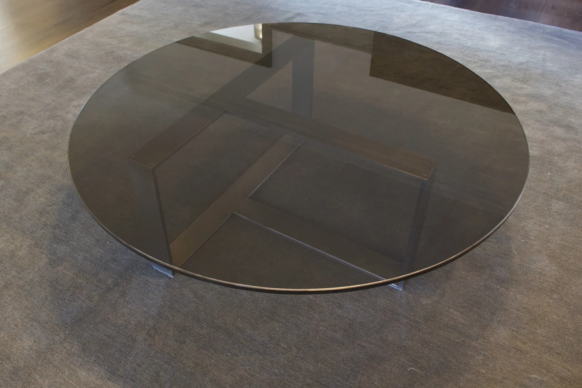 Minimalist round glass dining table with dark tinted top and metal base on polished concrete floor, designed by Adam Johnson.