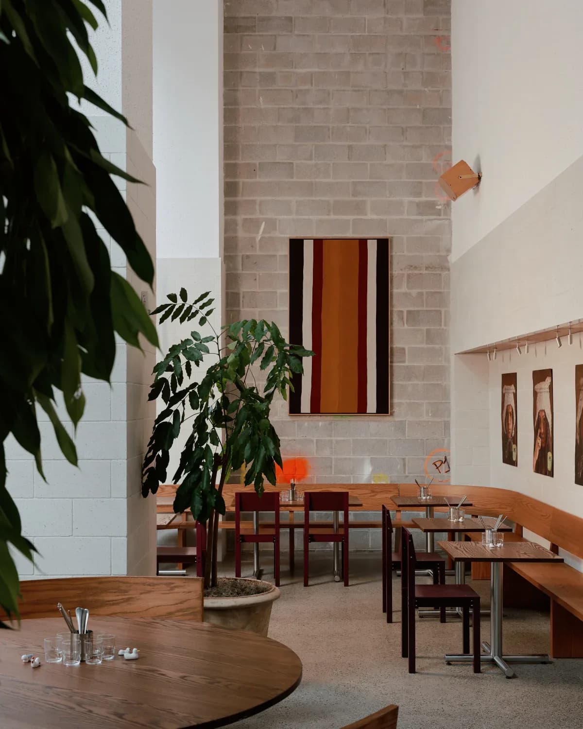 Modern dining room with brick walls, wooden tables and chairs, green plants, and abstract wall art in warm earth tones by Adam Rolston.