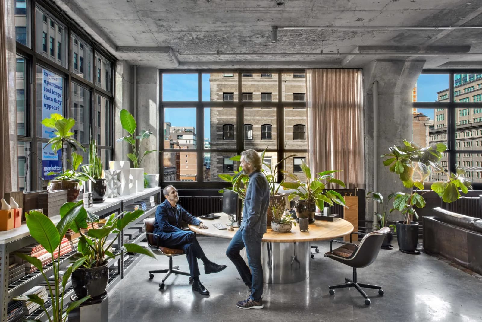 Modern loft office with concrete ceiling and floors, large windows overlooking NYC skyline, abundant greenery, wooden round table, and industrial-modern furnishings by Adam Rolston.