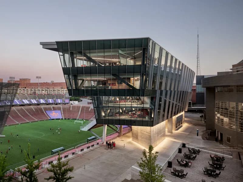 Modern dark-framed building with glass facade adjacent to a pink stadium, featuring illuminated courtyard with seating areas, designed by Adam Yarinsky.