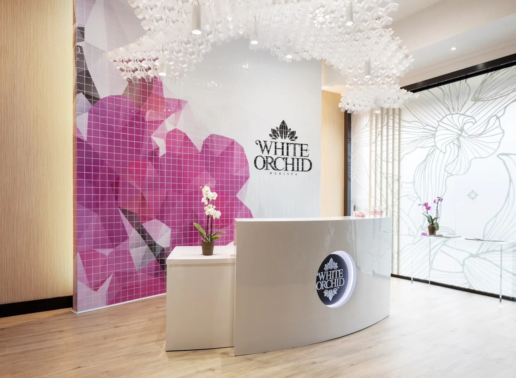 White Orchid spa reception area featuring a curved white desk, crystalline ceiling fixture, and striking magenta orchid-shaped wall mural by Adina Kornbluth.