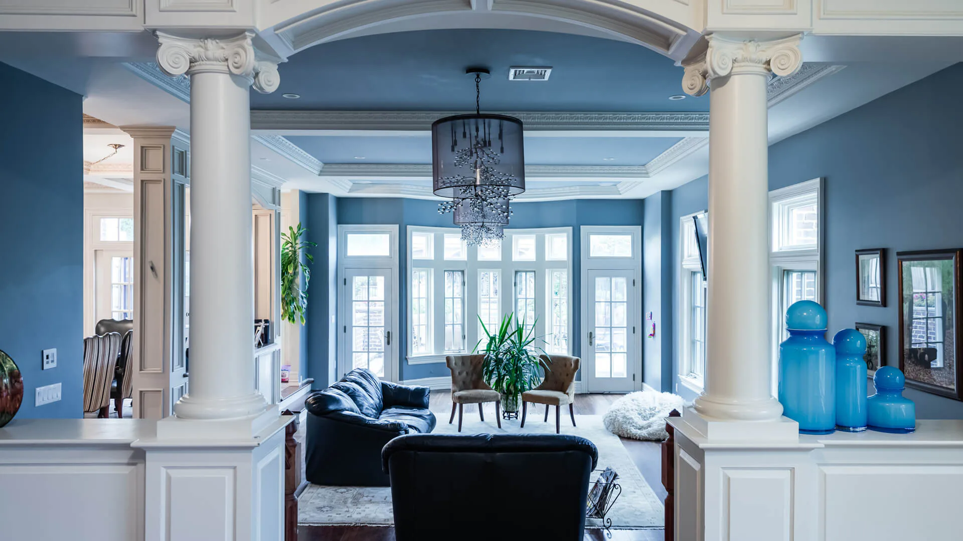 Blue-and-white foyer with classical columns, arched ceiling, glass doors, and pendant lighting, featuring a black sofa and potted plants by Adina Kornbluth.