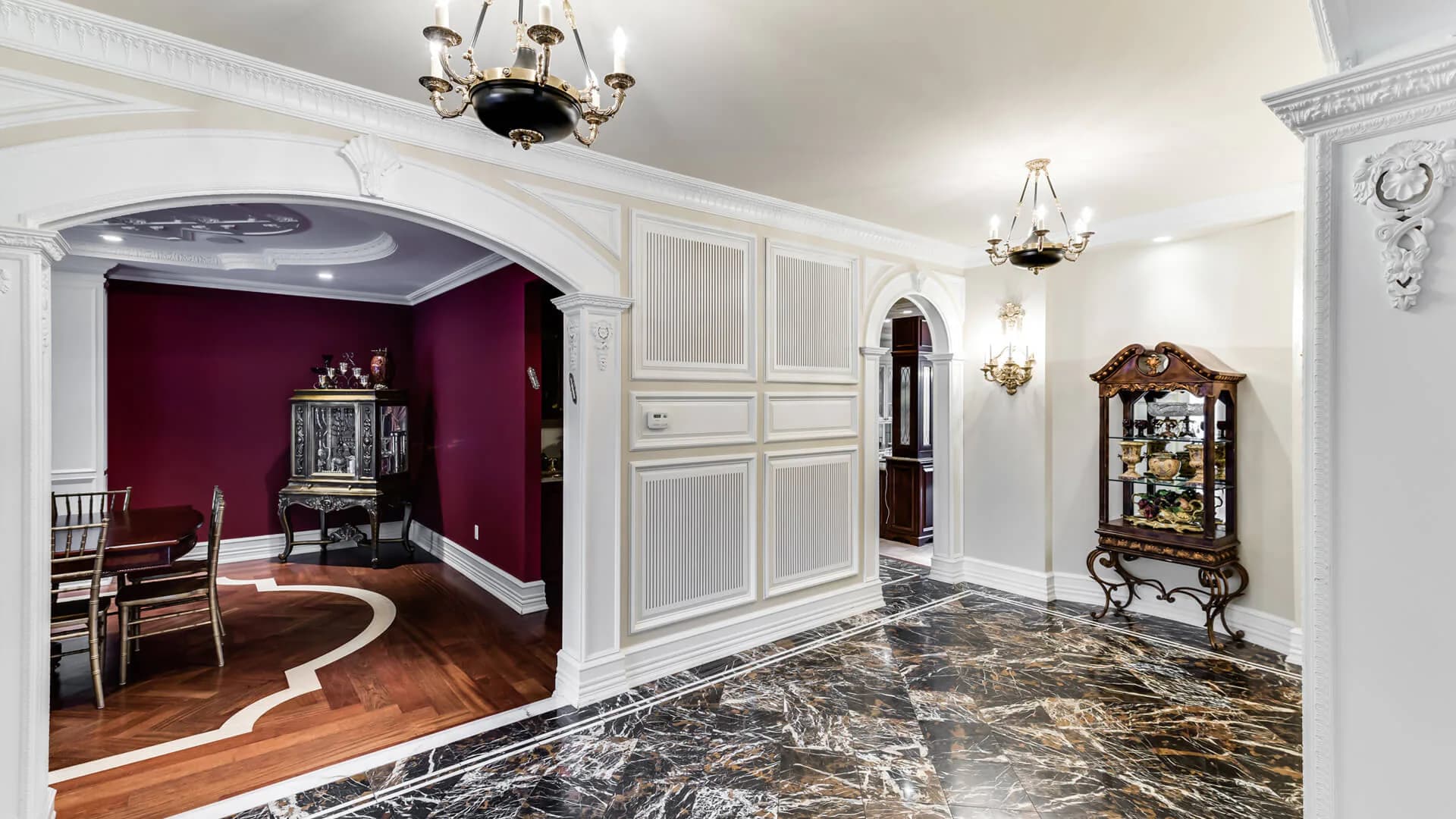 Luxurious foyer with marble flooring, white paneled walls, ornate chandelier, and burgundy velvet sitting room visible through archway, designed by Adina Kornbluth.