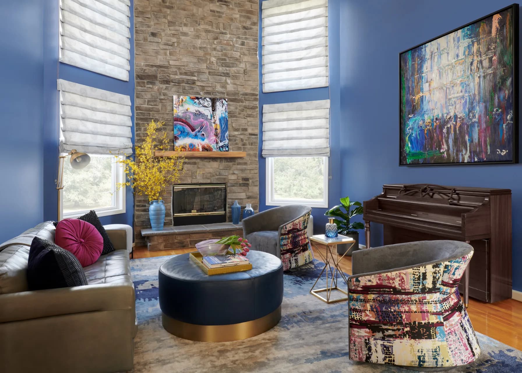 Modern living room with periwinkle blue walls, stone fireplace, round navy ottoman, colorful artwork, and wood piano by Adina Kornbluth.