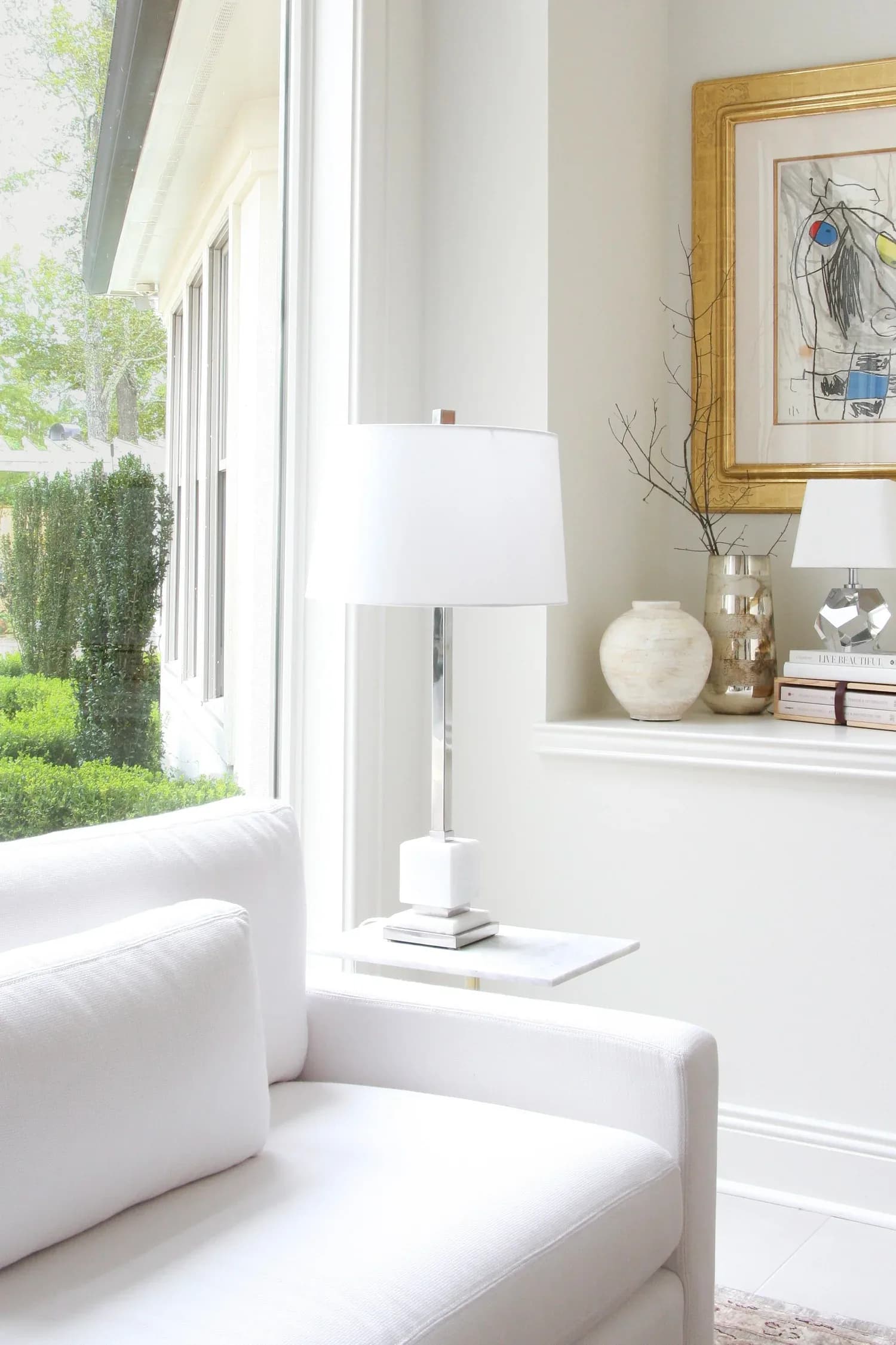 Bright, minimalist sitting area with cream upholstered chair, white table lamp, and styled shelf featuring gold-framed artwork and natural accents by Adrienne Jemison.