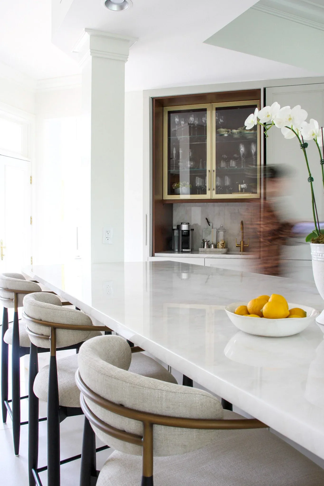 Modern kitchen with white waterfall-edge countertop, wooden cabinetry with glass doors, white bar seating with natural wood frames, and fresh lemons and orchid accents by Adrienne Jemison.