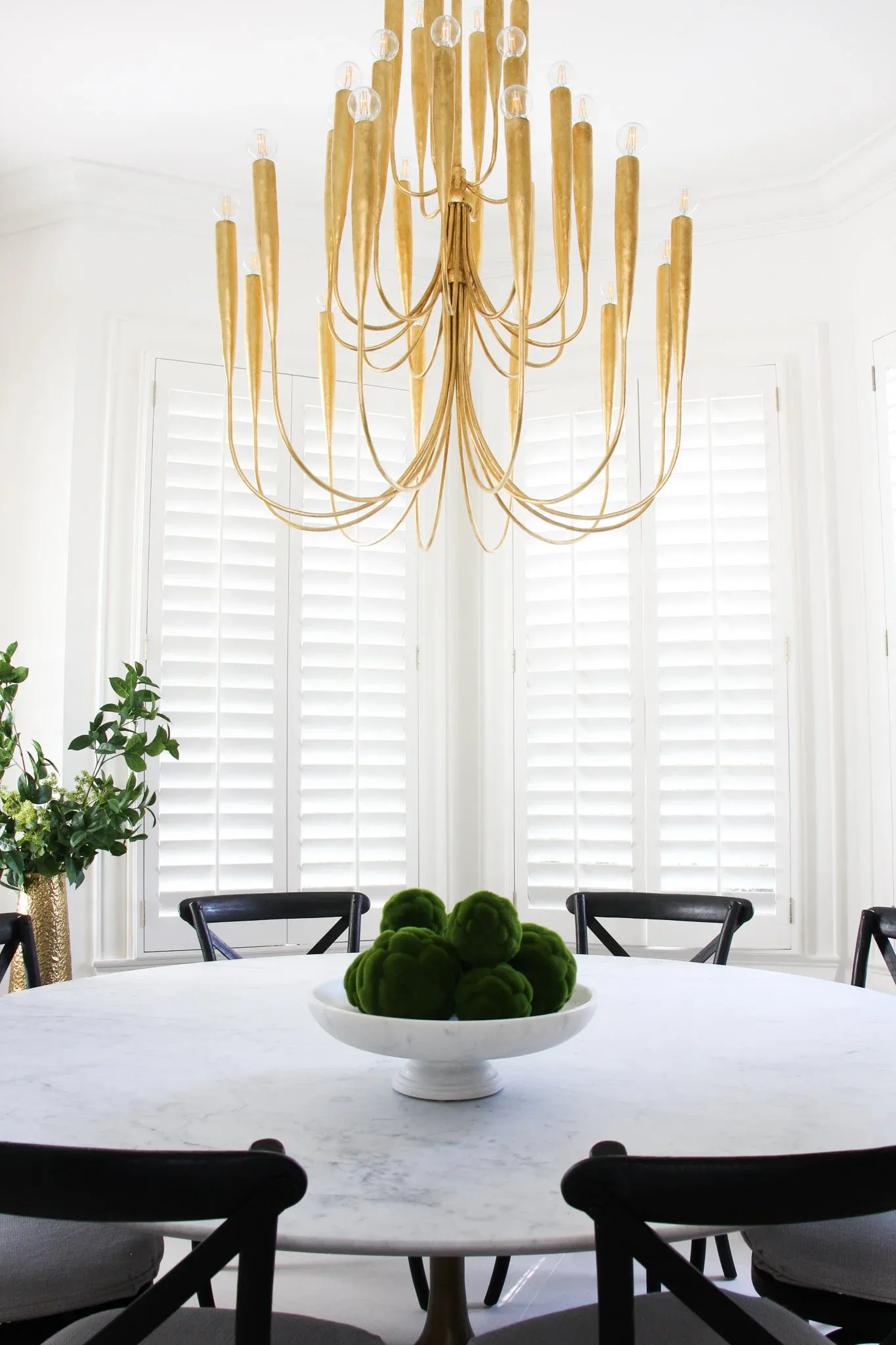A modern dining room features a white marble round table with black X-frame chairs beneath a sculptural brass chandelier, white shuttered windows, and potted greenery by Adrienne Jemison.