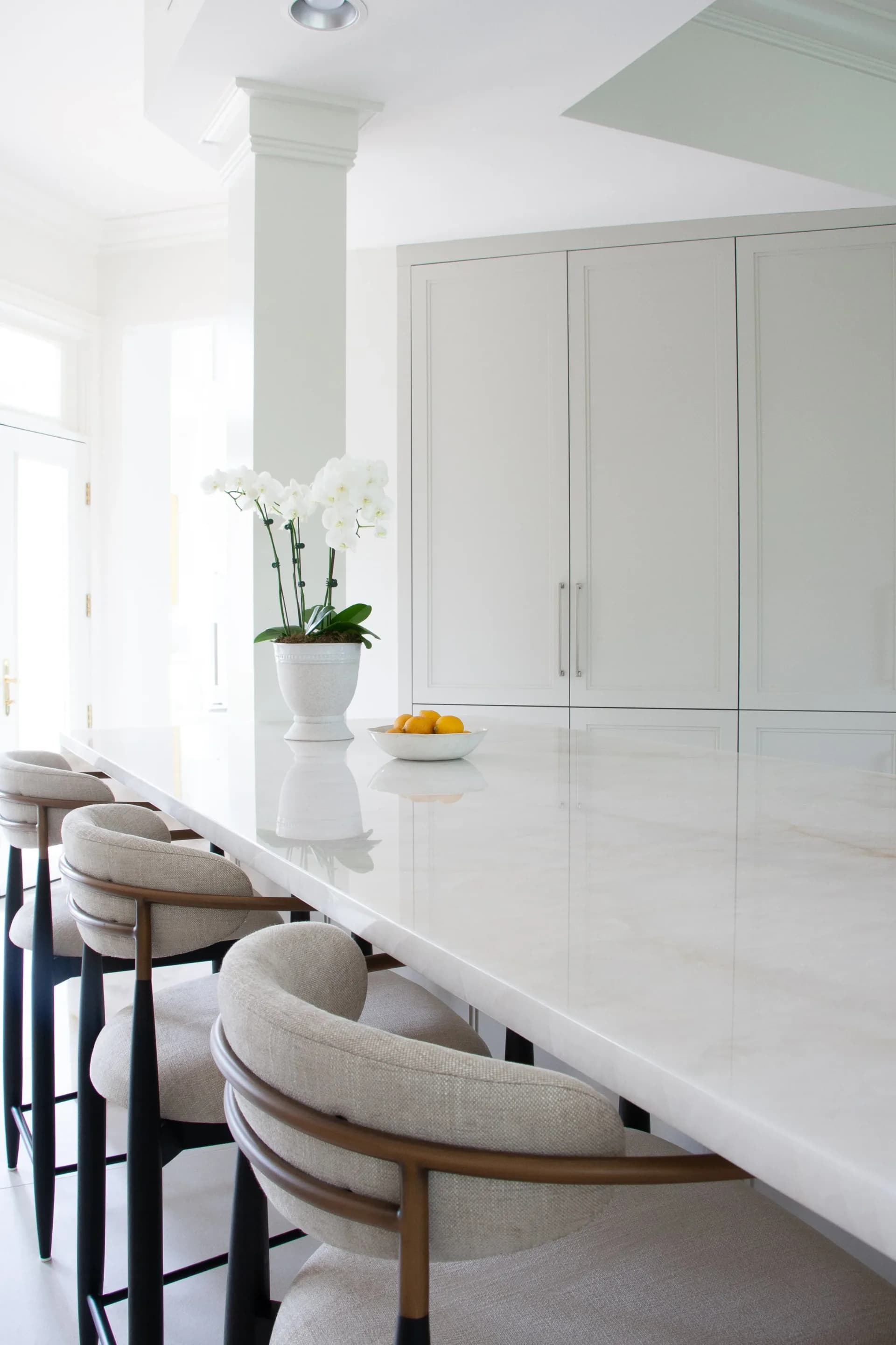 Minimalist white kitchen with glossy cabinetry, white marble waterfall countertop, wooden bar stools, orchid arrangement, and natural light from windows, designed by Adrienne Jemison.