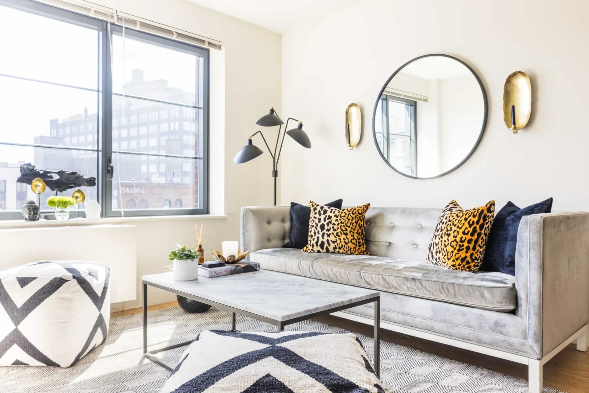 Bright living room with gray sofa, navy and gold leopard pillows, round mirror, geometric rug, and city views by Agata Zajkowski.