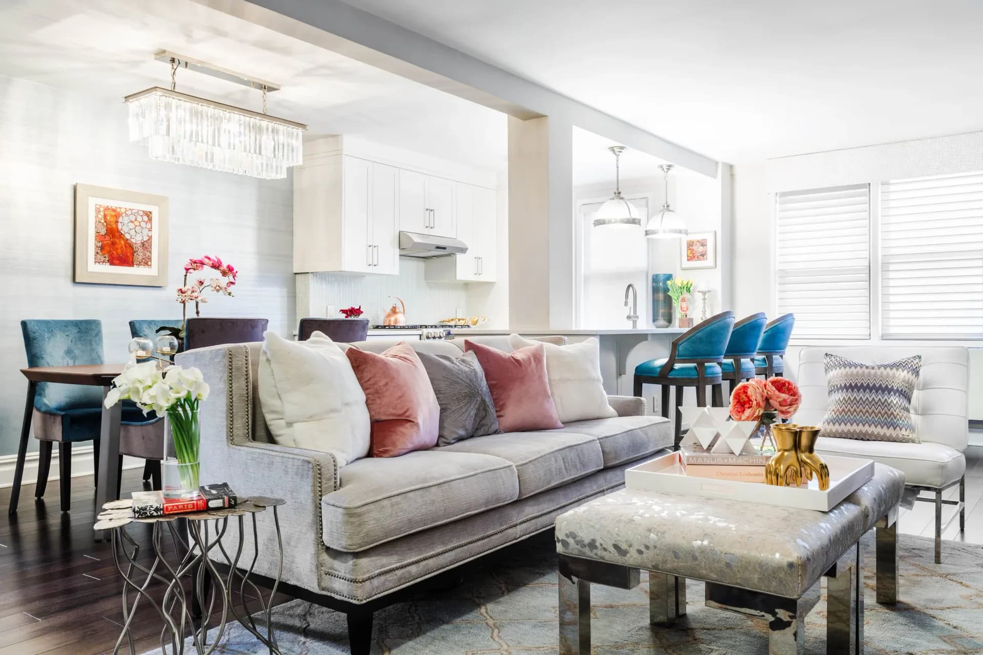 Open-concept living room with gray sectional sofa, colorful throw pillows, cream ottoman, blue accent chairs, and kitchen visible in background, designed by Agata Zajkowski.