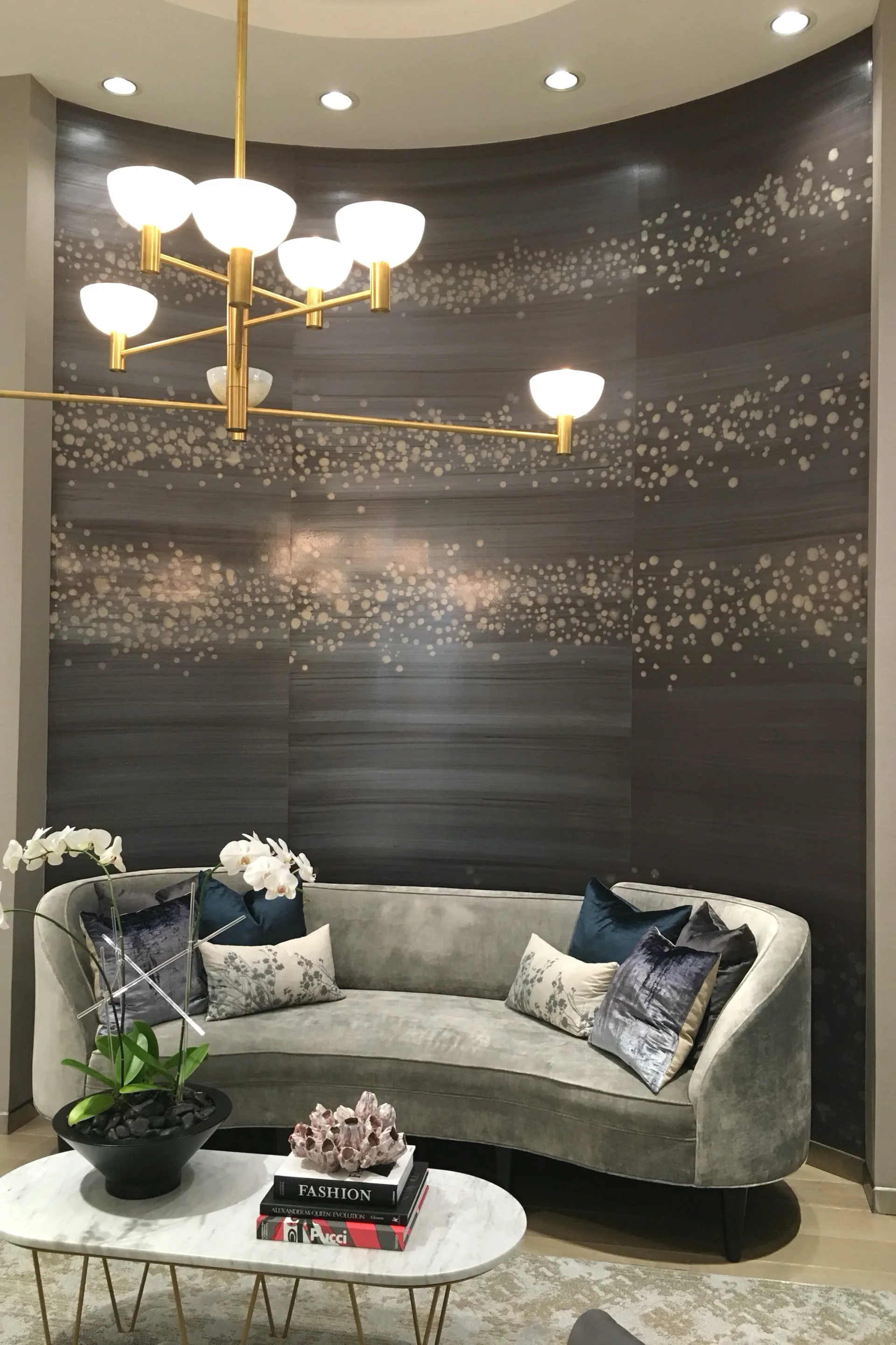 Curved gray upholstered sofa in a niche with dark textured wall backdrop and gold chandelier by Agnes Liptak.