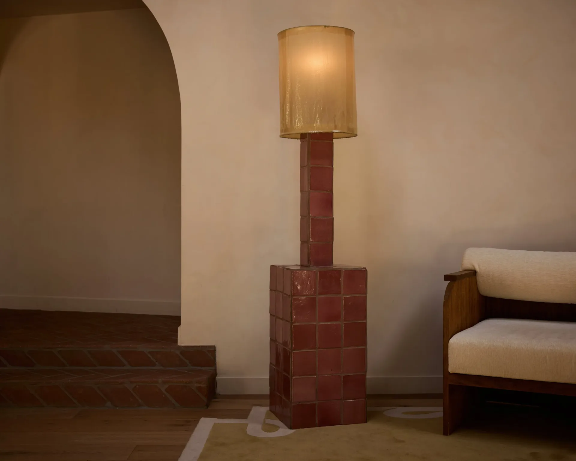 Minimalist interior with burgundy tile cylindrical pedestal supporting a cream linen floor lamp, concrete stairs, and cream seating, warm neutral palette by Ah Um Design Studio.