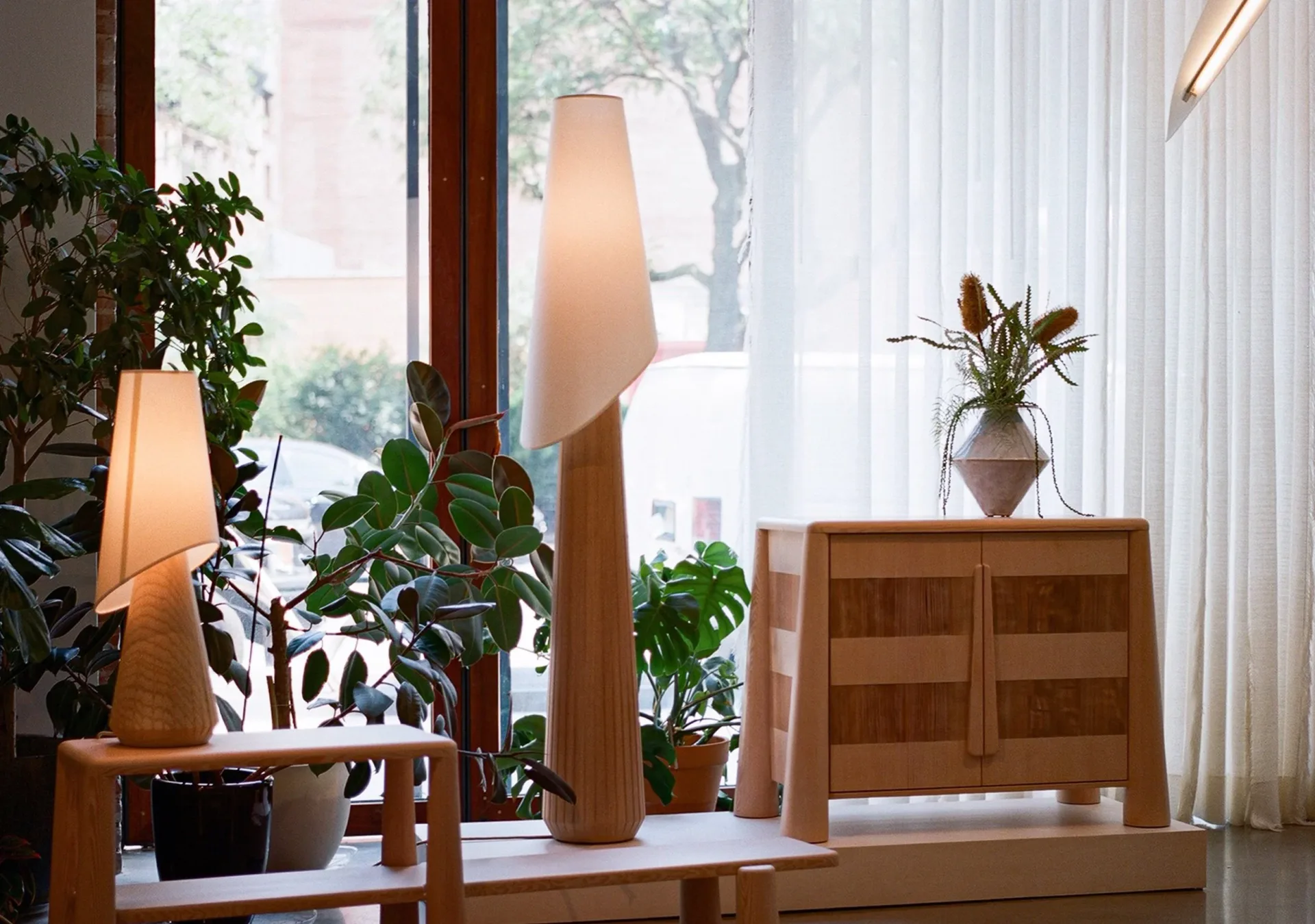 Minimalist interior with wooden furniture, potted plants, and sculptural floor lamps beside floor-to-ceiling windows by Alara Alkan.