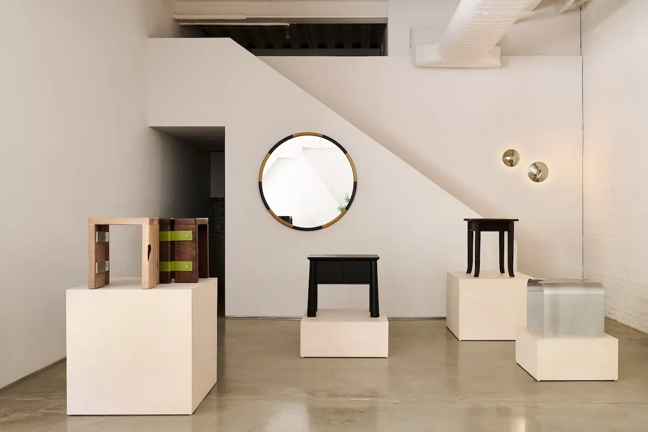 Minimalist gallery installation with white cubic pedestals, black stool, circular mirror, and sculptural objects on polished concrete floor by Alara Alkan.