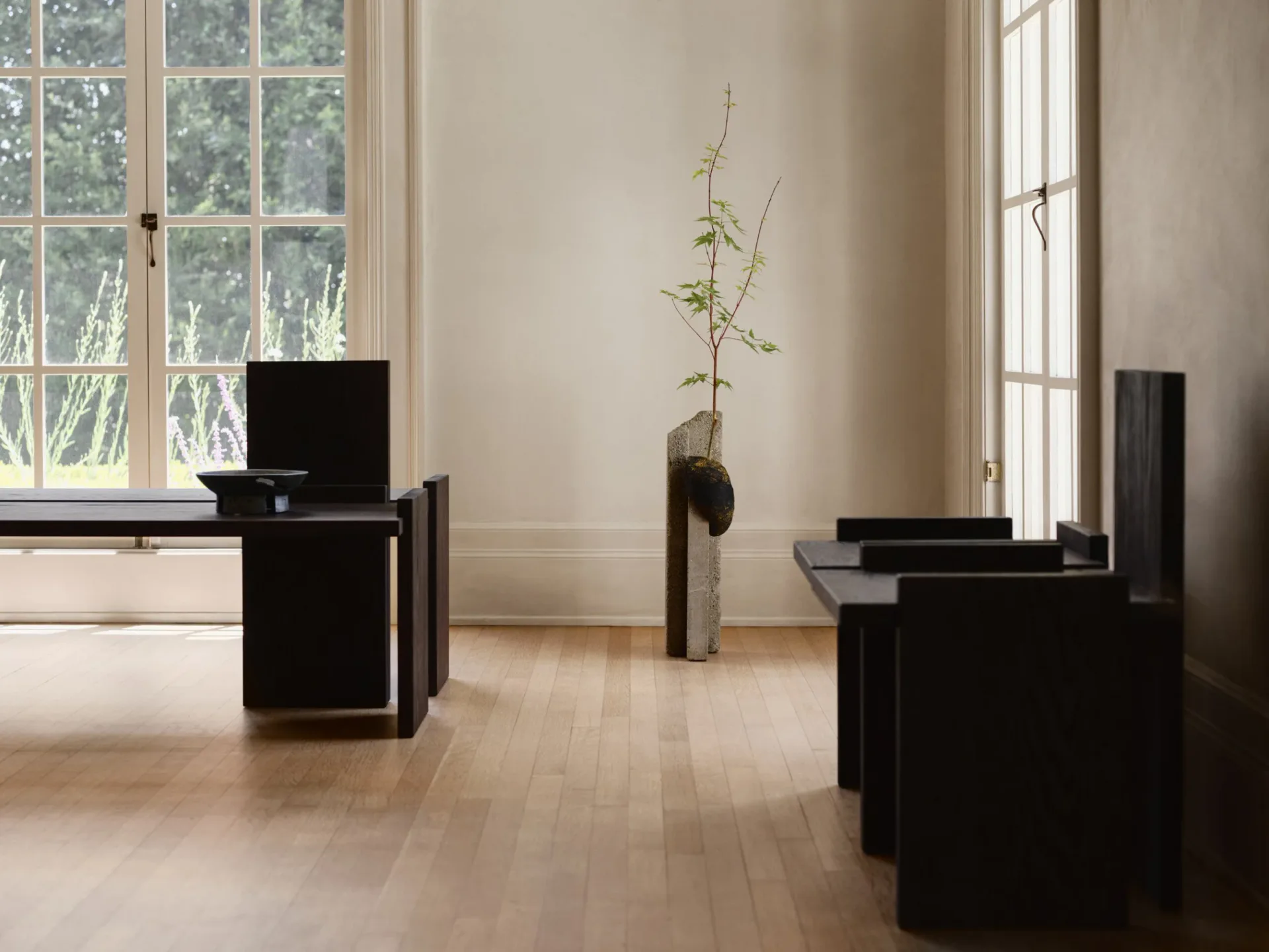Minimalist interior with light wood flooring, black furniture pieces, and large windows; a single delicate plant specimen in a vase anchors the serene, gallery-like space by Alejandro Bataille.