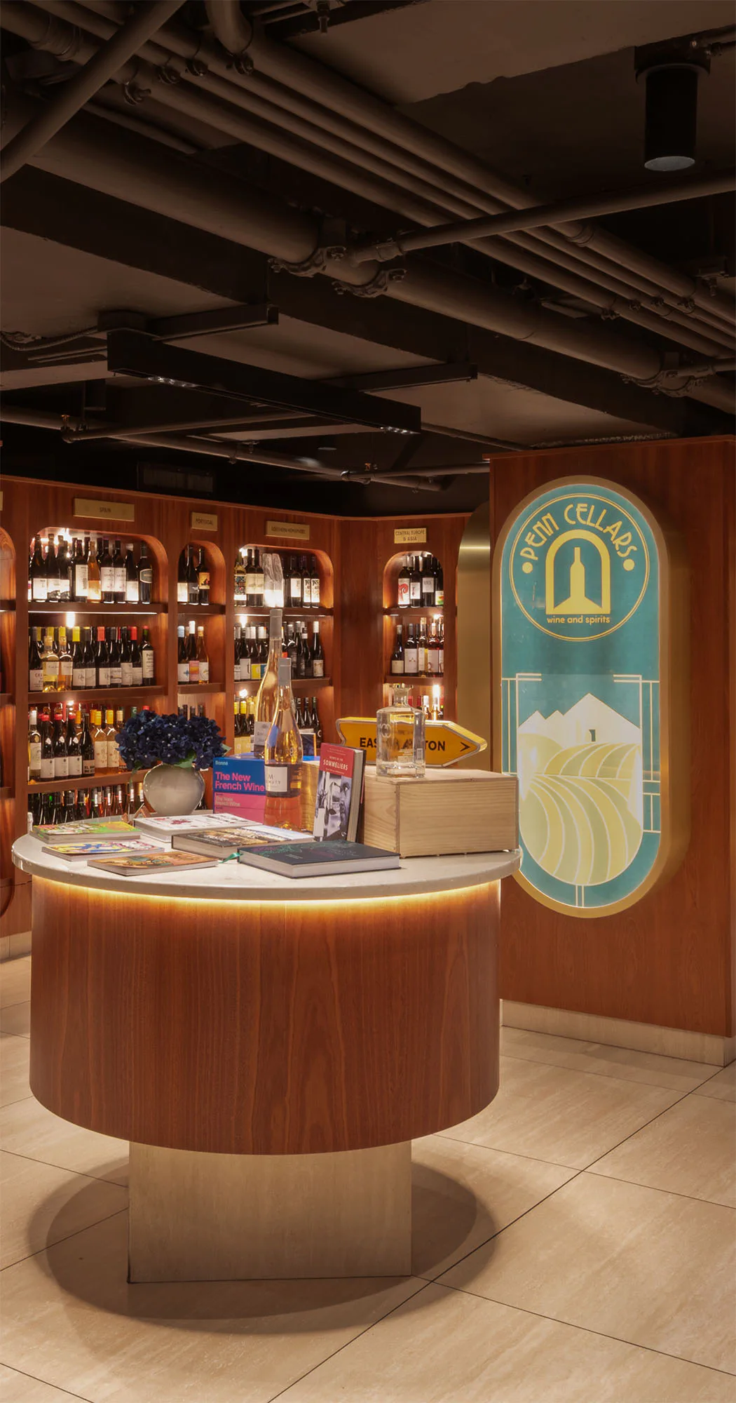 Warm wood-paneled retail counter with curved front, illuminated base, and shelving displaying bottles behind, featuring a teal brand logo signage by Alessandro Preda.