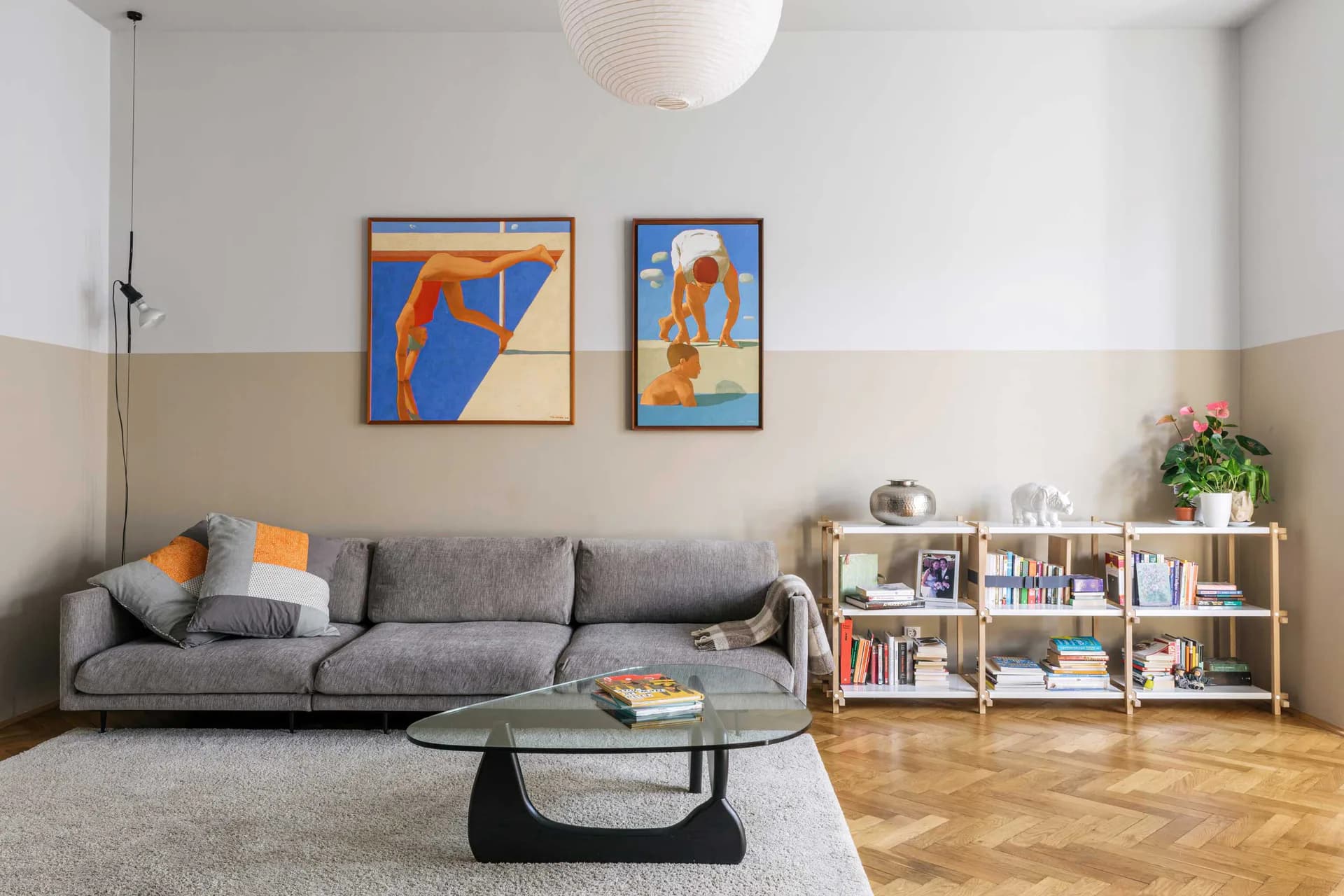 Modern living room with gray sofa, geometric artwork, two-tone wall treatment, wooden herringbone flooring, and white shelving unit by Alessandro Preda.