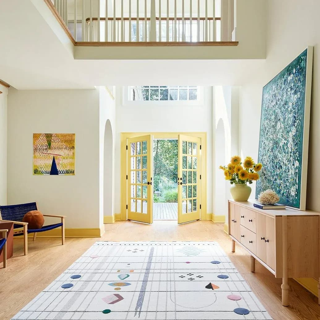 Bright, minimalist foyer with yellow double doors, wooden floors, patterned area rug, natural light, and mezzanine railing by Alex Barker.
