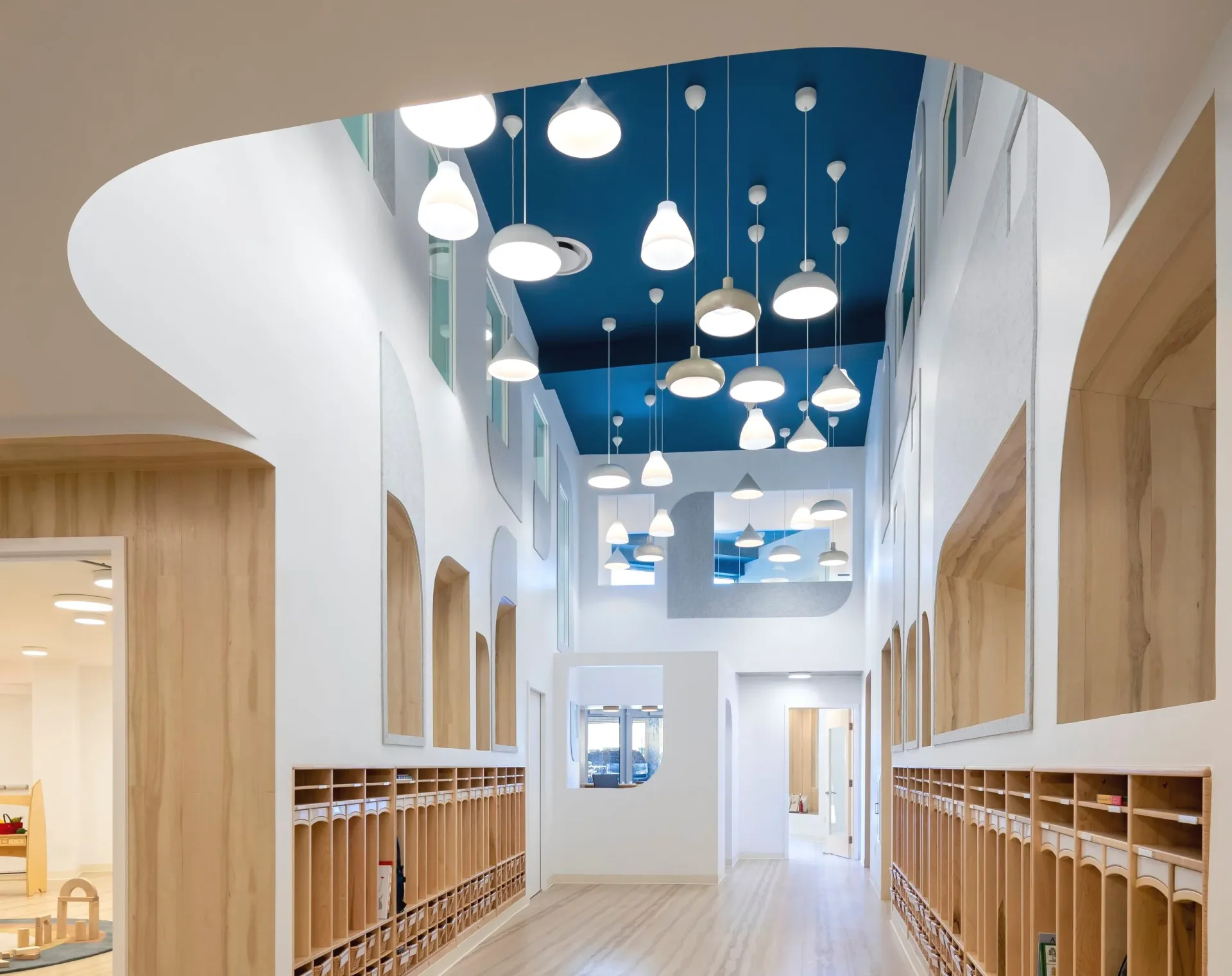 Modern pediatric clinic hallway with natural wood lockers, white walls, and suspended pendant lighting fixtures against a blue ceiling by Alex Barker.