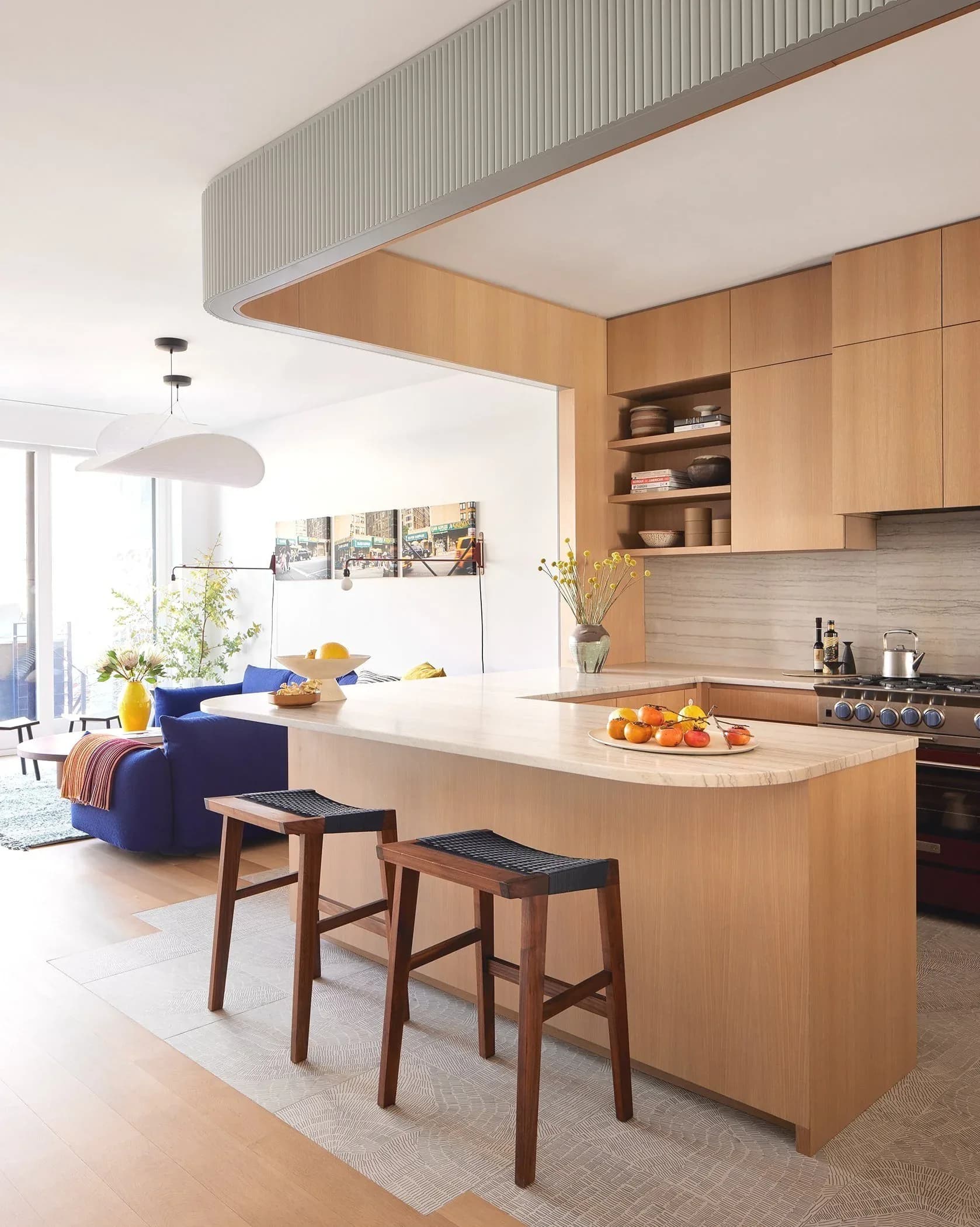 Modern open-plan kitchen and living space with light wood cabinetry, white countertops, natural materials, and wood bar seating by Alex Barker.