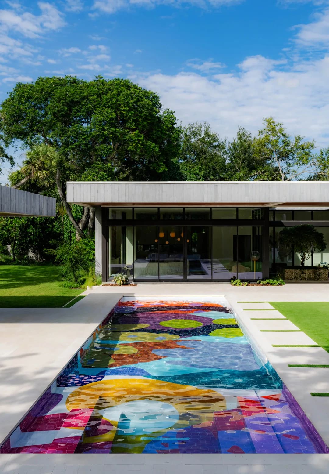 Modern residential courtyard features a vibrant multicolored geometric rug on concrete paving, with minimalist architecture and mature trees by Alex Proba.