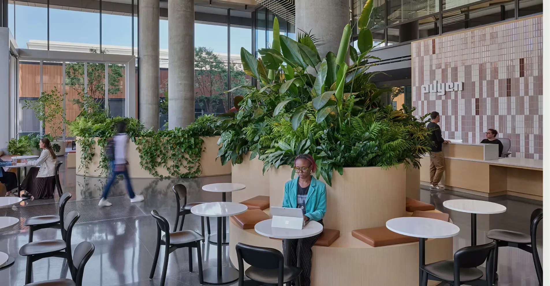 Modern office café space with large planted containers, round white tables, black chairs, and floor-to-ceiling windows overlooking a courtyard by Alexa Boyd.