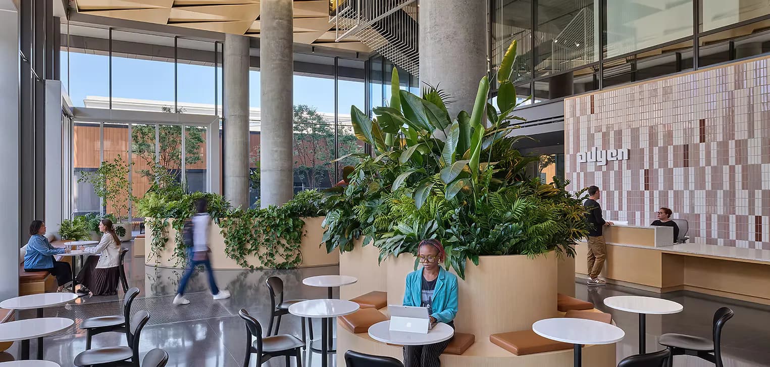 Modern office cafeteria with concrete columns, abundant green plants in large planters, mixed seating areas, and floor-to-ceiling windows overlooking autumn foliage, designed by Alexa Boyd.