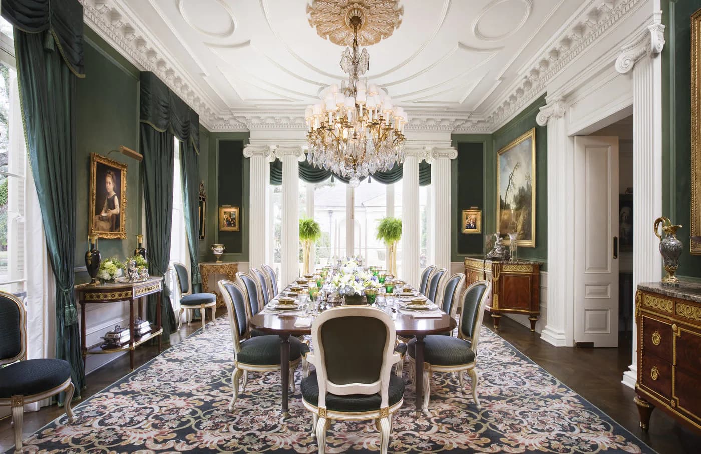 Formal dining room with deep green walls, ornate white ceiling medallion, crystal chandelier, and long table set with striped chairs by Alexa Hampton.