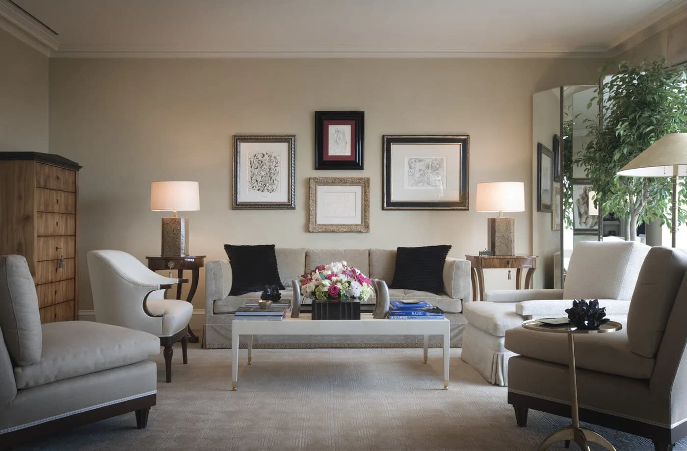 A sophisticated living room with neutral palette features two light gray upholstered chairs flanking a whitewashed coffee table, framed artwork above, table lamps, and modern furnishings by Alexa Hampton.