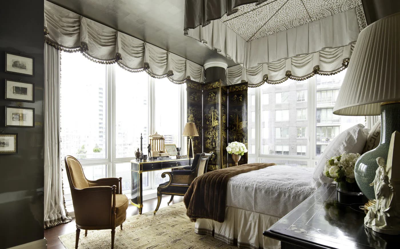 Luxury bedroom with dark walls, cream drapery with swags, four-poster bed, upholstered seating, and large windows overlooking city skyline by Alexa Hampton.