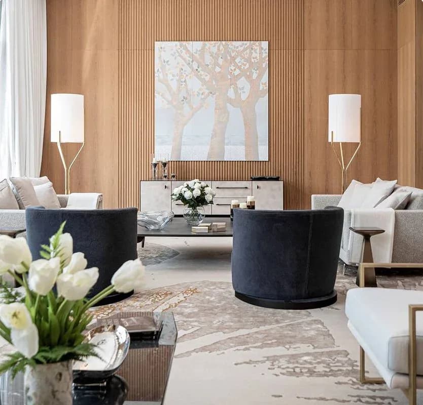Luxury living room with warm wood-paneled walls, two dark circular velvet chairs, neutral seating, and marble flooring by Alexandra Champalimaud.