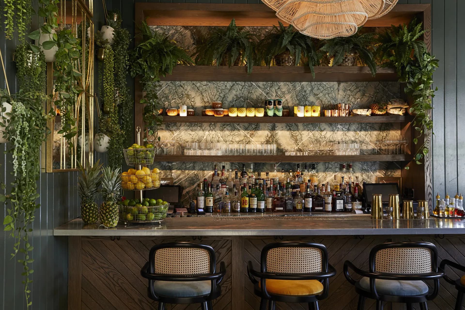 A sophisticated bar interior featuring dark wood cabinetry, backlit shelving displaying bottles, abundant greenery, brass accents, and three modern caned chairs, designed by Alexandra Champalimaud.