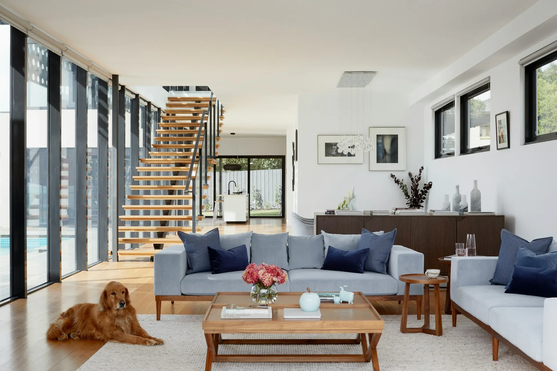 Bright contemporary living room with blue-upholstered sofas, wooden coffee table, striped curtains, and windows overlooking stairs, designed by Alexandra Grosser.