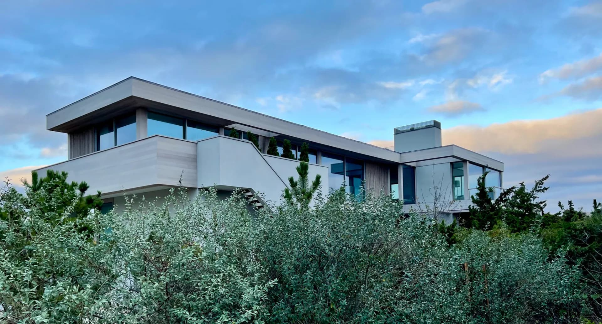 Modern residential architecture with clean lines and horizontal windows set above olive trees, featuring light concrete and metal cladding by Alexi Simmons.
