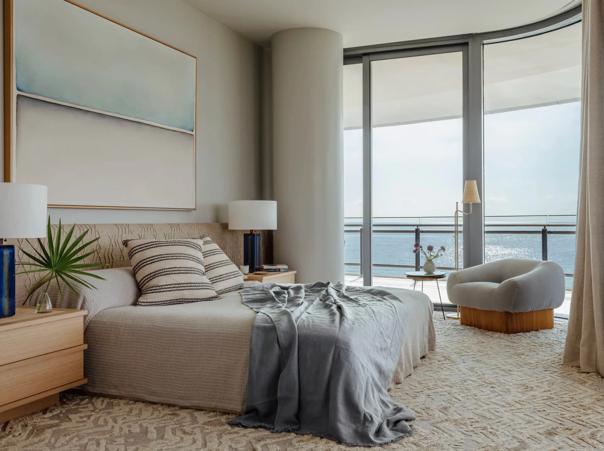 Waterfront bedroom with neutral palette, light wood furniture, striped pillows, ocean views through floor-to-ceiling glass doors, and curved accent chair by Alexi Simmons.