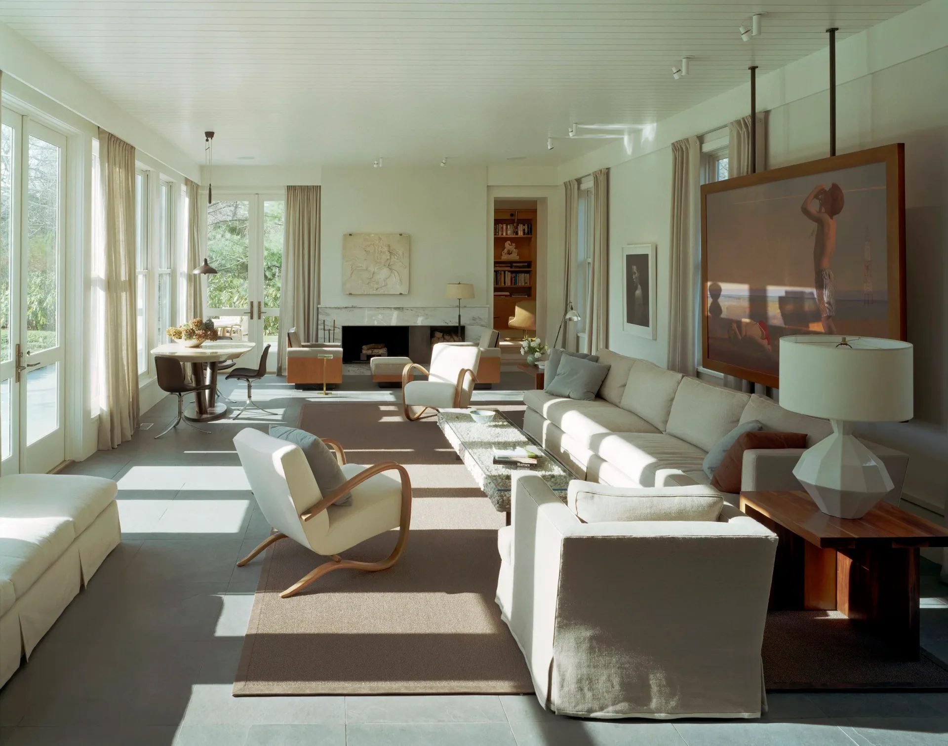 Spacious modern living room with cream and white furnishings, dark wood flooring, large windows, fireplace, and mid-century modern accents by Alexi Simmons.