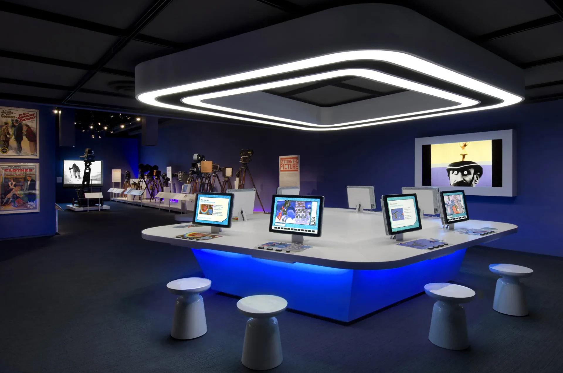 Modern exhibition space with blue walls, geometric lighting fixture, and multiple display screens arranged on a central platform with seating by Ali C. Höcek.
