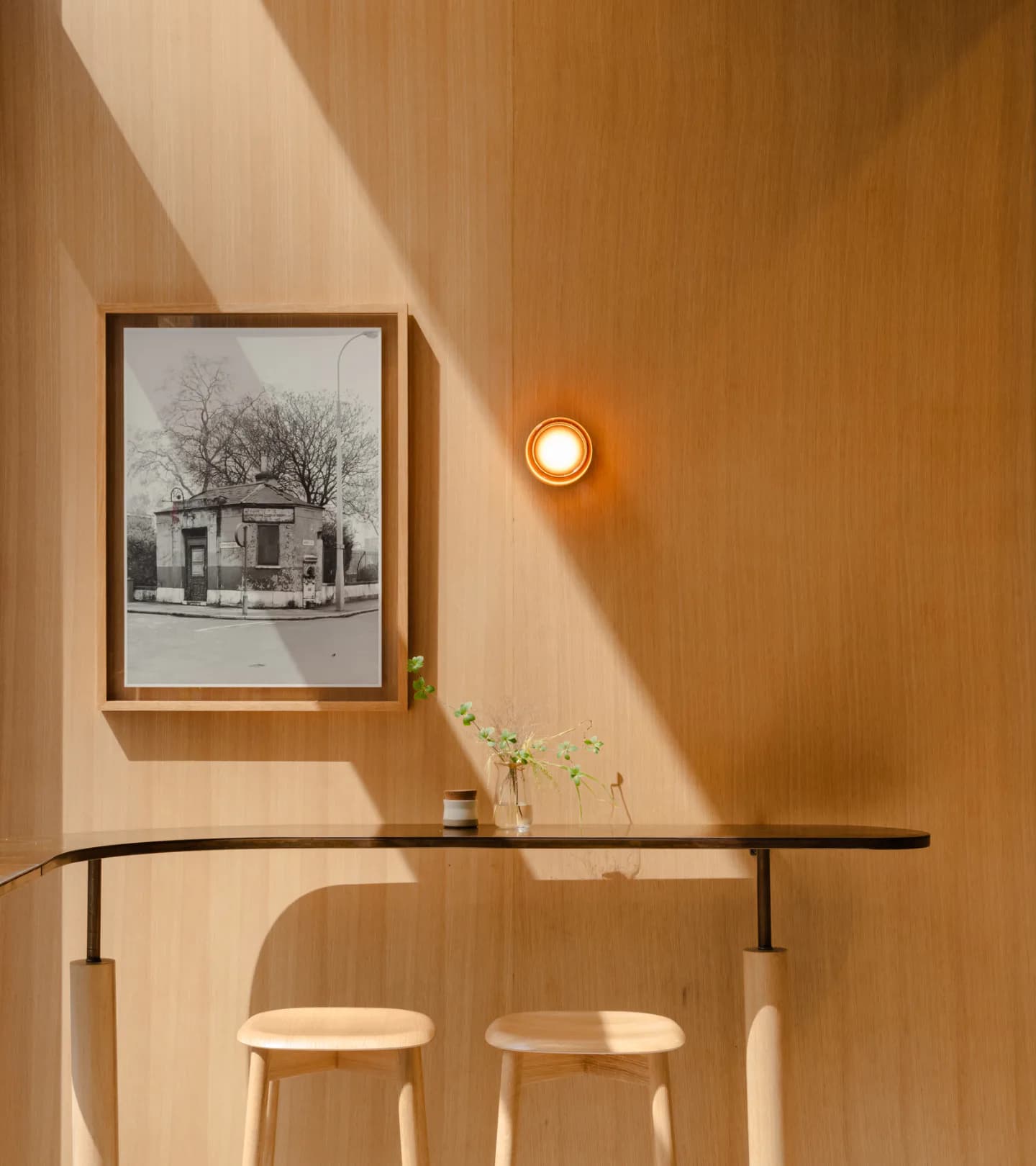 Minimalist interior with warm wood-paneled walls, a black console table with two cream stools, framed black-and-white photograph, recessed lighting, and fresh flowers by Alia Marzuki.