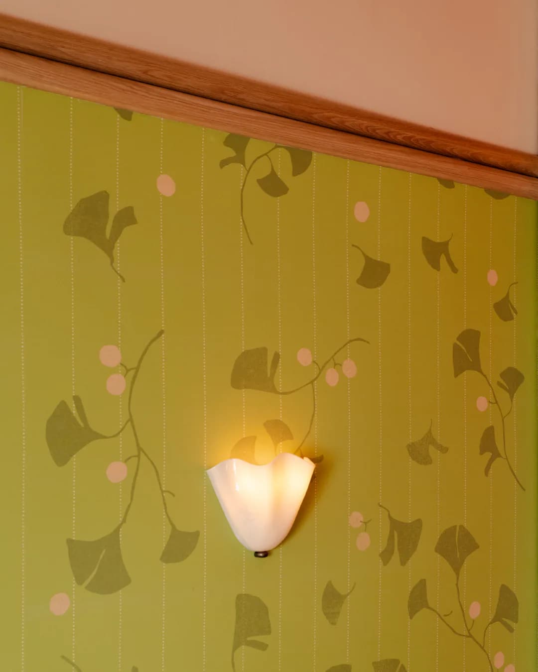 Modern wall sconce with frosted glass shade mounted on yellow-green wallpaper featuring botanical leaf and berry motifs, by Alia Marzuki.