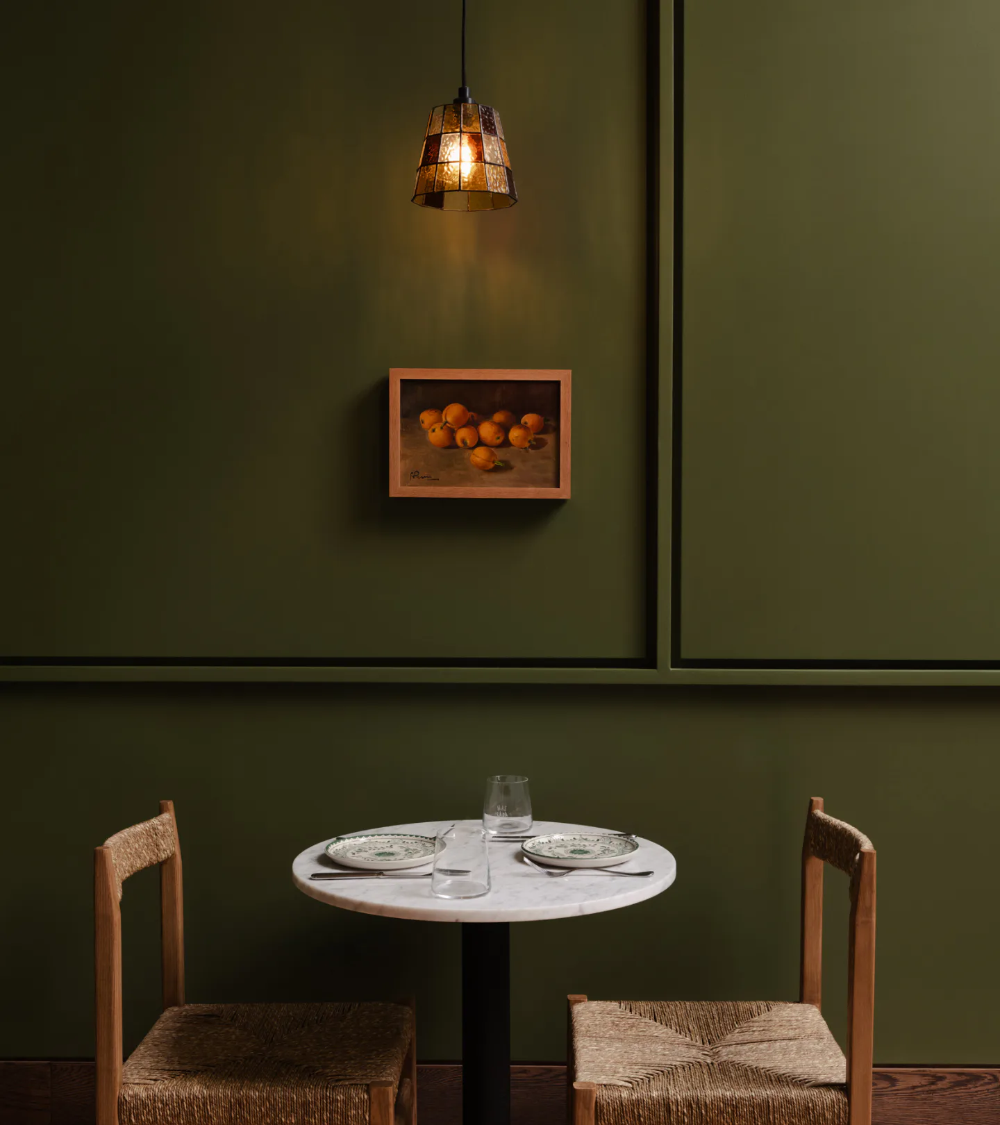 Minimalist dining space with sage green walls, round white table, woven-seat wooden chairs, pendant lighting, and framed orange artwork by Alia Marzuki.